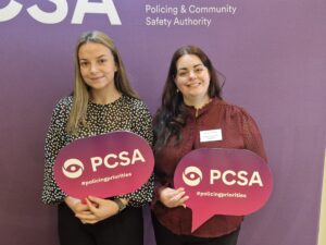 Two women hold signs in front of a purple banner