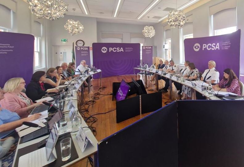 A meeting room with people seated in a U-shape around tables, all looking at screens and papers. Purple PCSA banners on the walls.