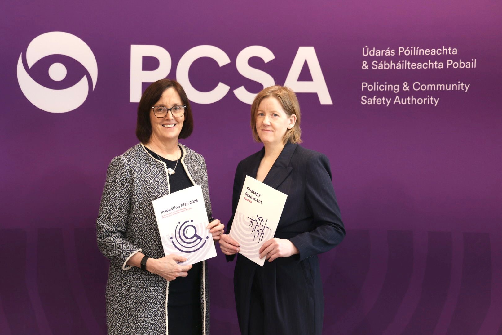 Two women stand in front of a purple banner holding two beige-covered reports