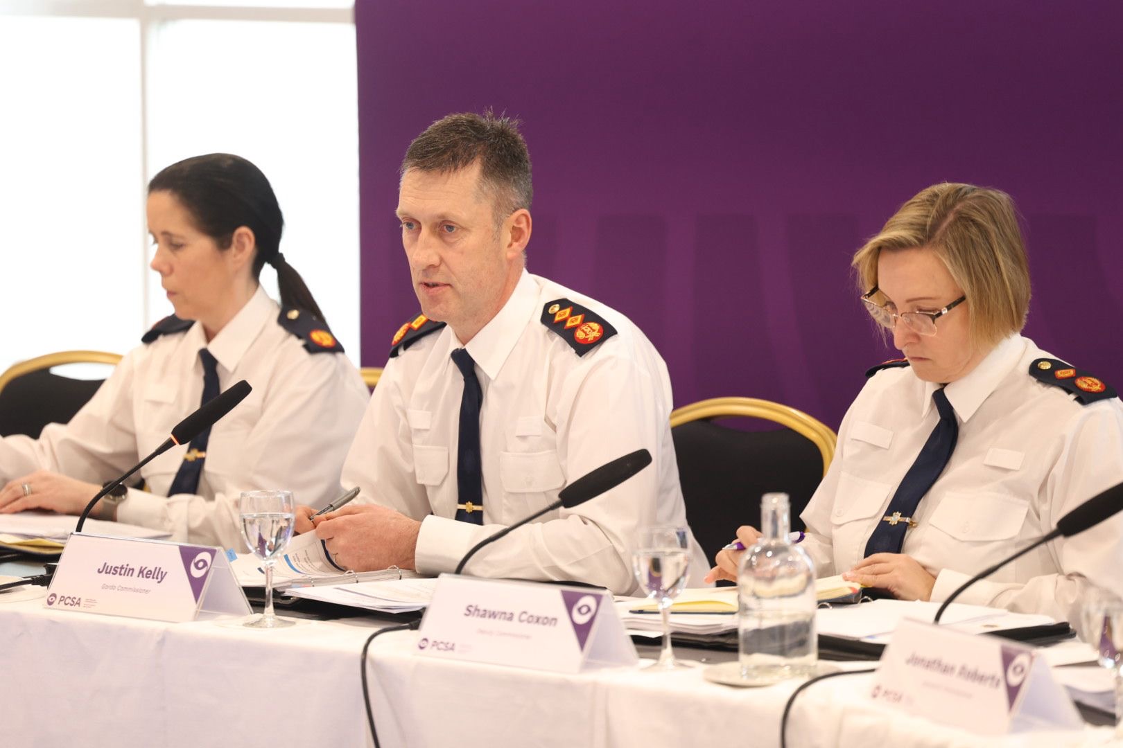 Garda Commissioner - Man in white short sits in front of desk and purple banner speaking