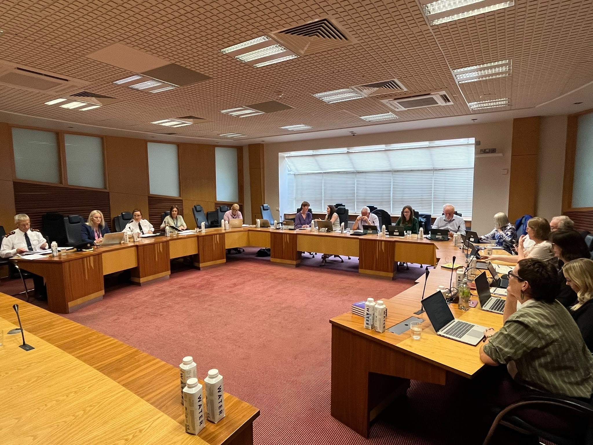 A large boardroom with a U-shaped arrangement of wooden conference tables. Around fifteen people are seated with laptops, documents, microphones, and water cartons in front of them. The room has a red carpet, wood-paneled walls, high-backed chairs, and large windows with closed blinds along one side. Ceiling lights brightly illuminate the meeting space as participants engage in a formal discussion.