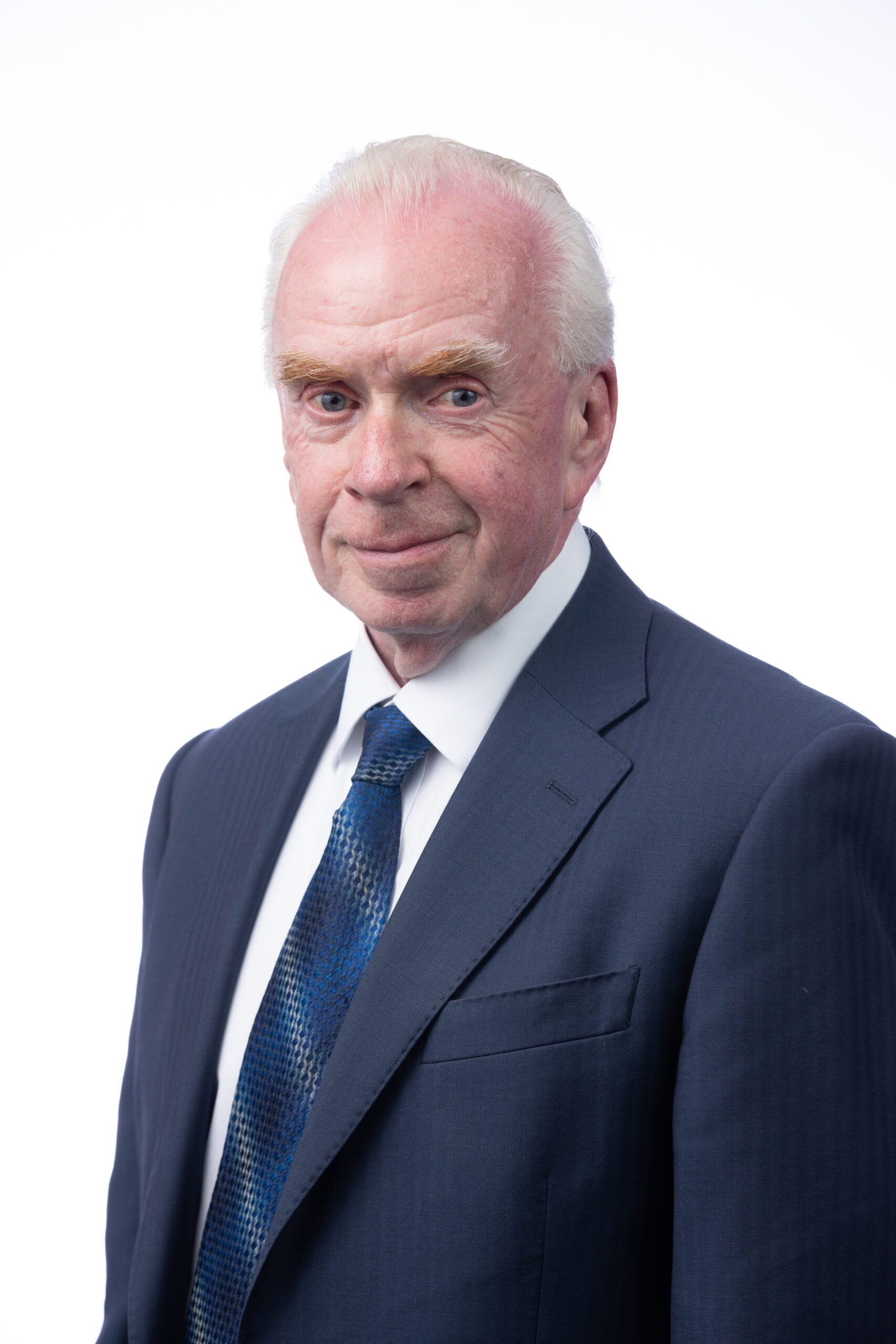 A man wearing a navy suit, white shirt and navy tie poses for an official photograph in front of a white background